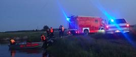 Pojazdy straży pożarnej stoją na brzegu rzeki. Strażacy wodują łódź