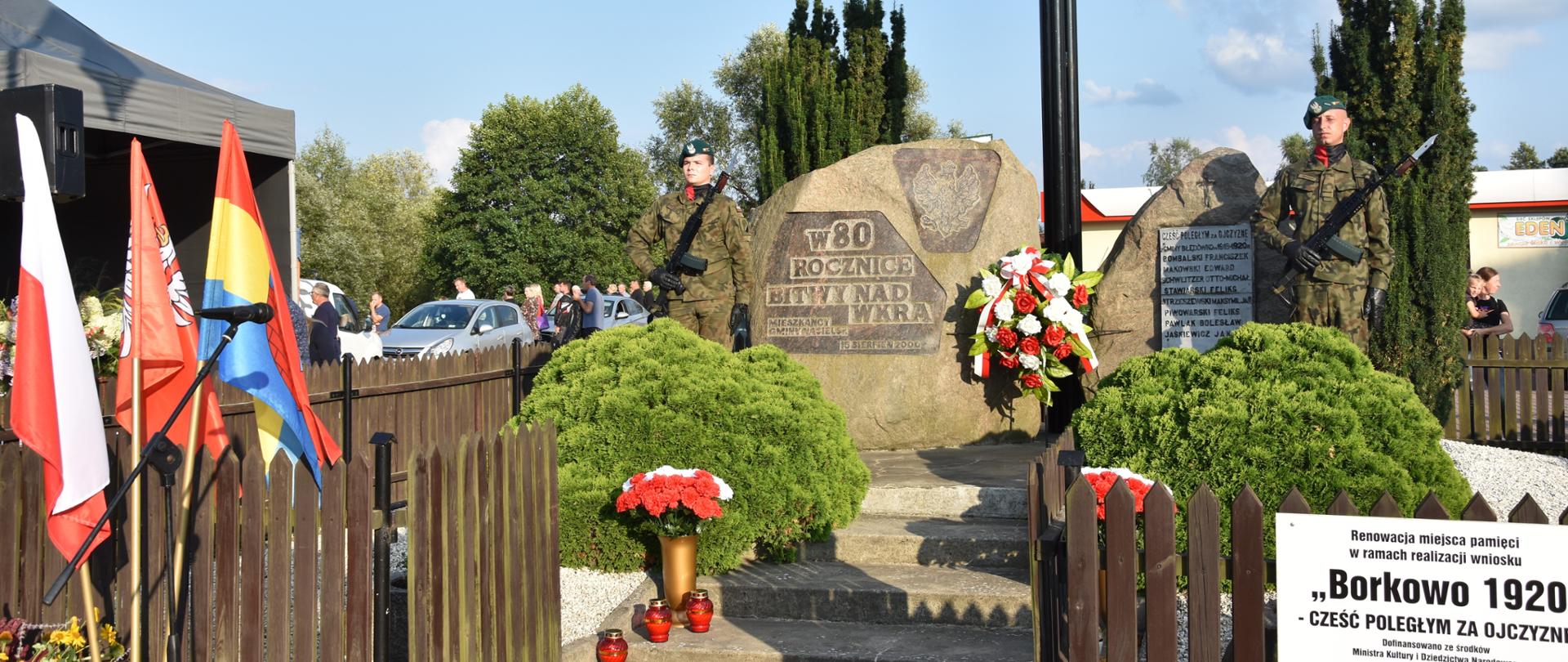 Zdjęcie przedstawia pomnik, upamiętniający Bitwę nad Wkrą oraz stojący przy nim wysoki krzyż. Przy pomniku asystę honorową pełnią żołnierze Wojska Polskiego. 