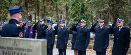 Po lewej stronie zdjęcia w pierwszym planie stoi półprofilem strażak z warty honorowej w wyjściowym mundurze. W głębi kadru, przodem do obiektywu, stoi szereg delegacji strażackiej salutującej przed cmentarną mogiłą. Na pierwszym planie po lewej stronie widoczny jest zabytkowy kamienny nagrobek z wyrytym napisem „BAGIEŃSKA”. Uczestnicy, w tym reprezentanci PSP, ZOSP RP i kapelani, ubrani są w ciemnogranatowe płaszcze służbowe oraz czapki rogatywki, a jeden z kapelanów ma na sobie fioletową stułę liturgiczną. Tło tworzy aleja cmentarna ze skąpanymi w jesiennych barwach liśćmi oraz rosnącymi wzdłuż niej drzewami.