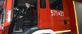 Za kierownicą wozu strażackiego siedzi druhna z ZHP. Ma na sobie ciemne spodnie i kurtkę. Na drzwiach od przedziału załogi znajduje się biały numer operacyjny 571P21. 