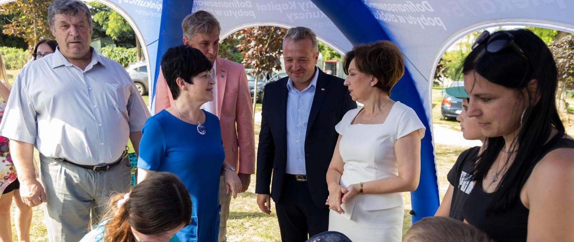 
Grupa ludzi - dorosłych i dzieci - stojąca na wolnym powietrzu
