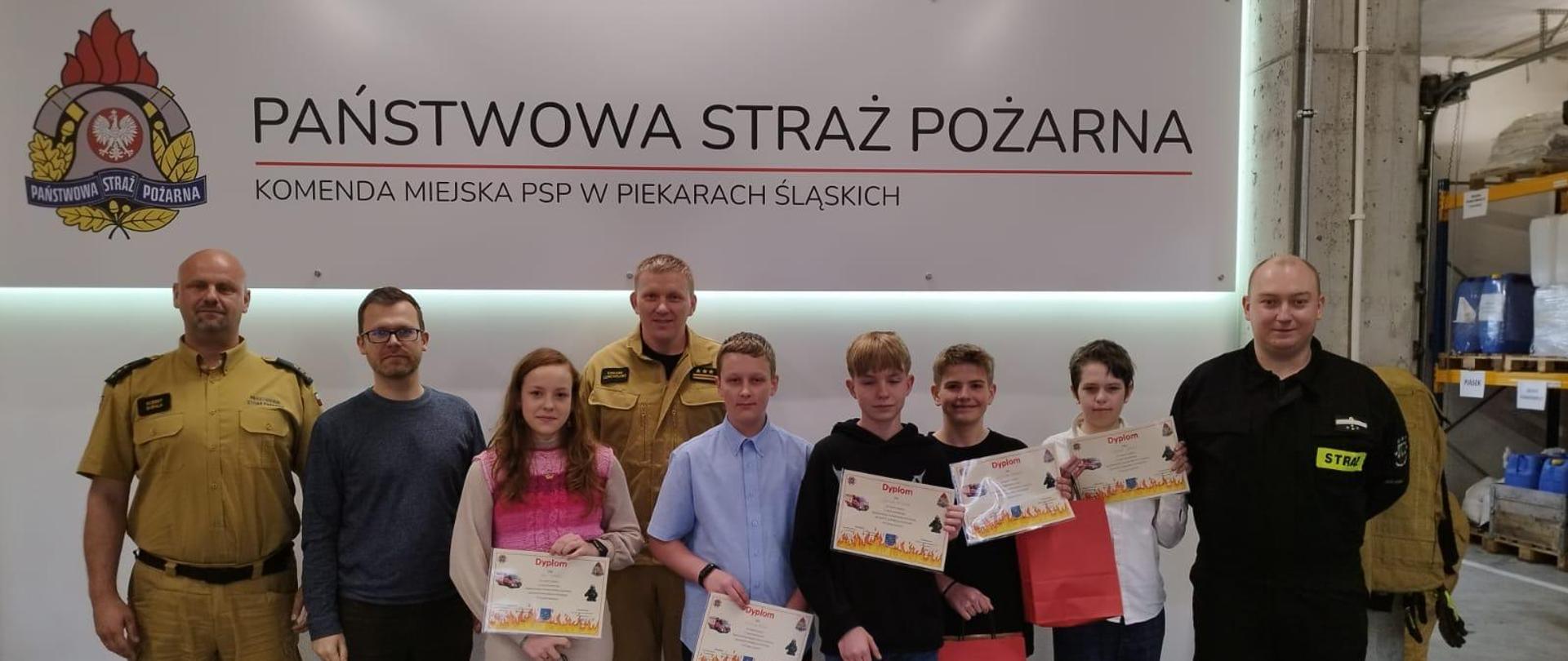 Na zdjęciu widać stojących w garażu przed banerem z logo PSP oraz nazwą komendy miejskiej w Piekarach Śląskich laureatów turnieju w kategorii wiekowej II w obecności Komendanta Miejskiego PSP w Piekarach Śląskich st. bryg. Tomasza Dziechciarza oraz Prezesa Zarządu Powiatowego Związku OSP RP w Piekarach Śląskich druha Marcina Matonia