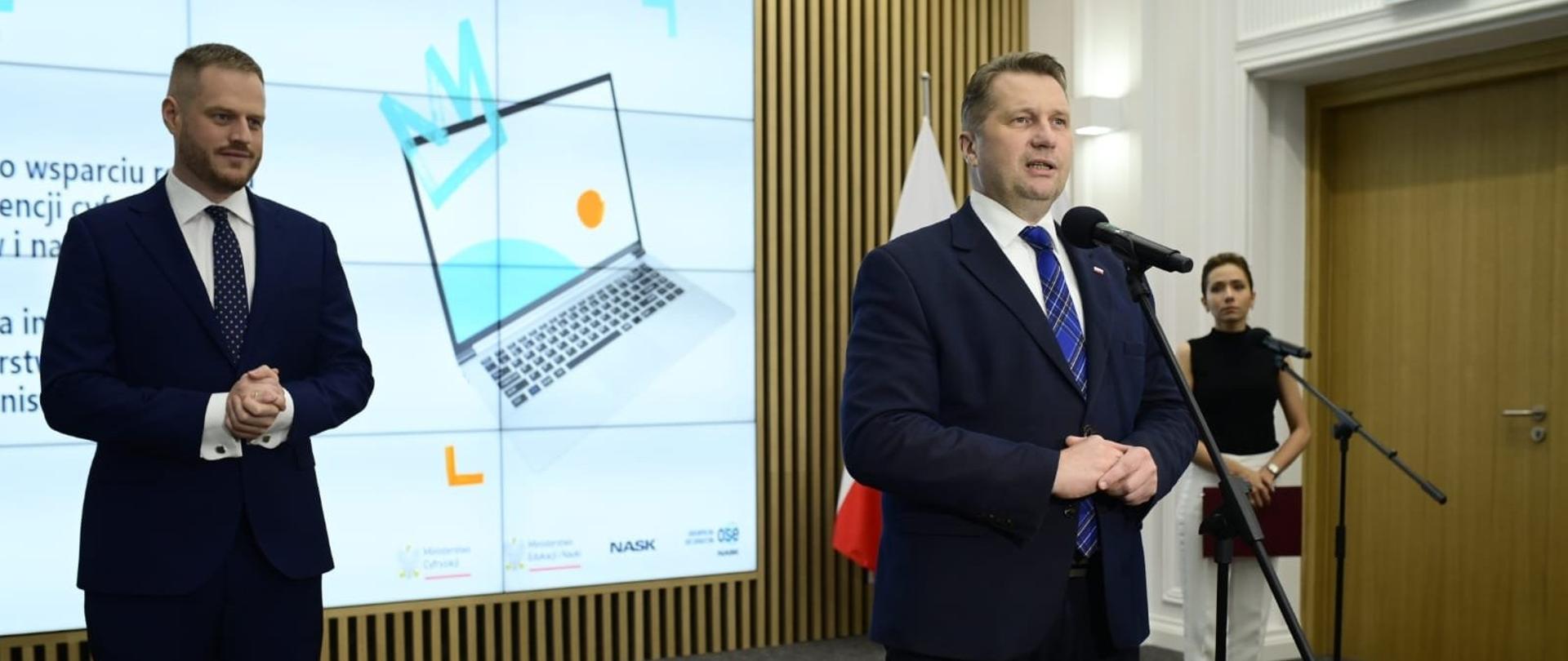 Minister Czarnek stoi przy mikrofonie obok niego po lewej mężczyzna w garniturze. W tle widać kobietę przy mikrofonie i telebim.