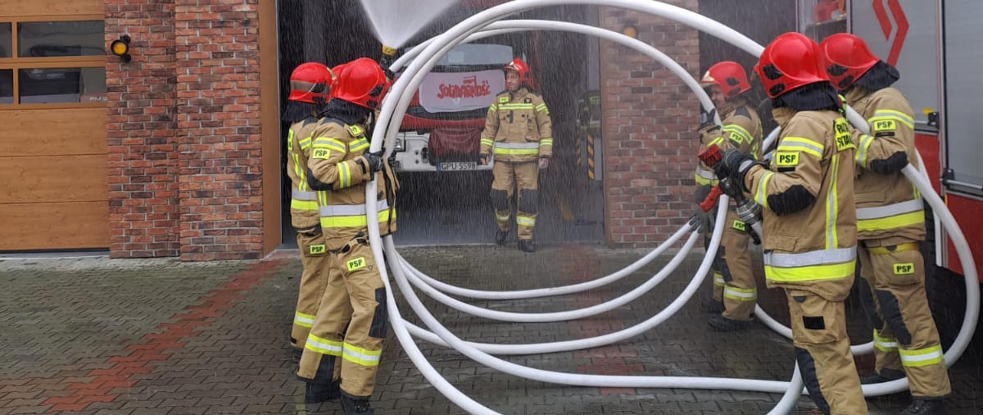 Na zdjęciu widać strażaków ubranych w ubrania specjalne, którzy trzymają wąż strażacki i polewają wodą swojego kolegę.