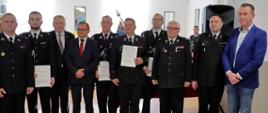 dziesięć osób pozuje do pamiątkowego zdjęcia trzy są ubrane w garnitur i marynarkę sześć jest ubranych w mundur wyjściowy ochotniczej straży pożarnej a jedna w mundur wyjściowy państwowej straży pożarnej cztery trzymają podpisane promesy w tle stolik z obrusem barwy czerwonej jasny parkiet na podłodze w tle sali dwa okna i lustra na ścianie