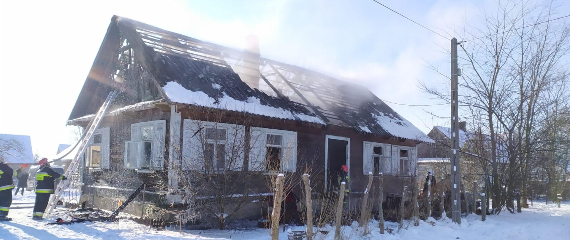 Widoczny śnieg, niska temperatura, zawalony dach drewnianego budynku, pora dzienna,widoczne węże pożarnicze, strażacy pracujący na drabinie w szczycie budynku oraz przy wejściu do budynku