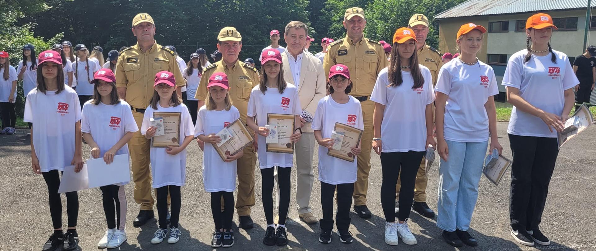Zdjęcie zrobione na zewnątrz, podczas uroczystej zbiórki. W szeregu stoją uczestnicy obozu szkoleniowo-wypoczynkowego młodzieżowych drużyn pożarniczych, razem z nimi stoi oficerowie Państwowej Straży Pożarnej.
