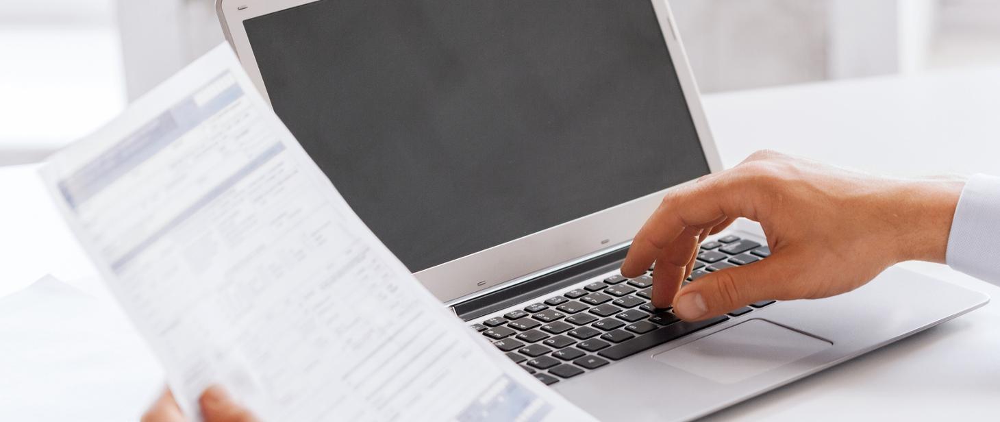 Laptop na biurku oraz jedna dłoń trzyma sprawozdania a druga dotyka klawiatury komputera