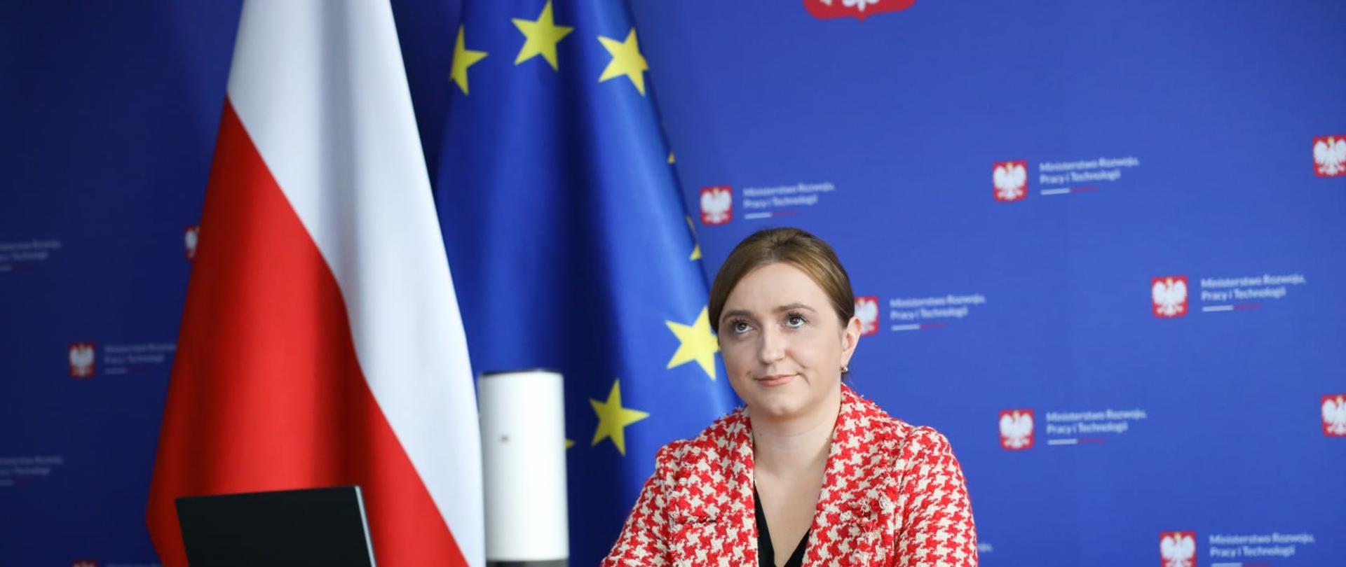 Wiceminister Olga Semeniuk podczas konferencji „Kadry Przyszłości - Kadry dla Przemysłu”. Wiceminister siedzi przy stole, po prawej stronie ma flagę Polski i Unii Europejskiej, z tyłu banek ministerstwa rozwoju. 