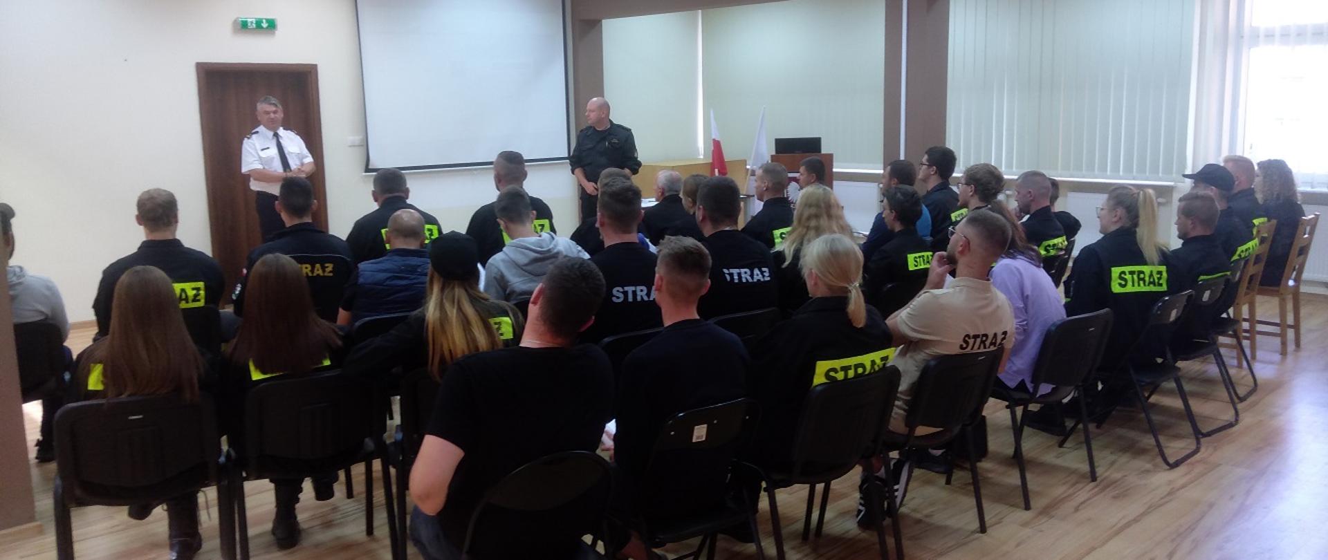 Zdjęcie przedstawia strażaków ochotników oraz funkcjonariuszy PSP podczas rozpoczęcia szkolenia podstawowego OSP.
W tle pomieszczenie świetlicy w budynku Komendy.
