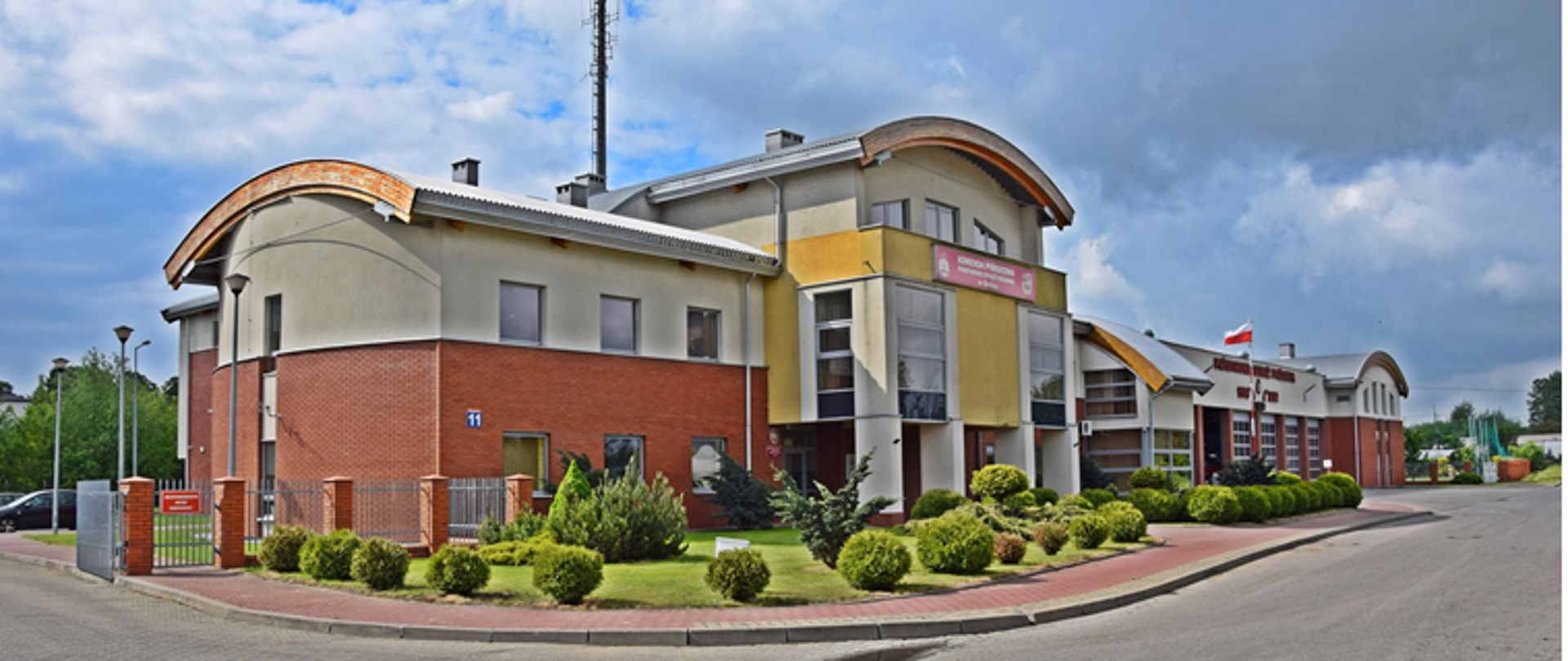 Zdjęcie przedstawia budynek Komendy widziany z przodu. Na zdjęciu widoczny jest maszt z flagą państwową, napis Państwowa Straż Pożarna, maszt antenowy. Budynek w kolorach czerwonej cegły oraz odcieniach żółtego. Przed budynkiem tereny zielone z krzewami. 