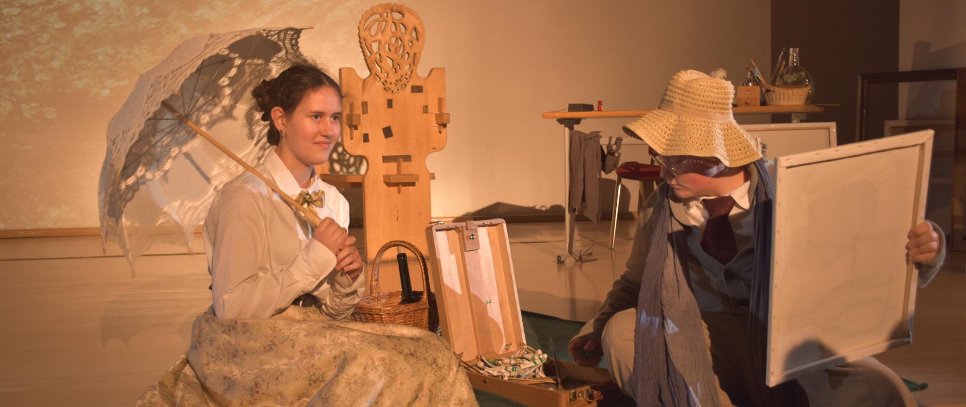Dwoje aktorów - kobieta ubrana w stroju z XIX wieku z parasolką w dłoni pozująca malarzowi w kapeluszu, który tworzy swój obraz.