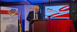 Chargé d’affaires Jacek Chodorowicz przemawia na na Global Gateway Forum 2026 w Maputo; w tle dwa roll-upy, w tym roll-up Polskiej Izby Handlowej; na drugim napis jest niewidoczny; w środku flaga mozambicka; z prawej strony ekran, na którym wyświetlony jest napis: Encontro B2B com Empresas da Polonia. Ekran stoi na stole przesłoniętym czerwonym materiałem