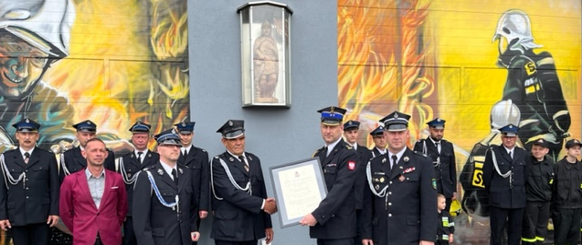 Na zdjęciu widoczne uroczyste przekazanie aktu włączenia jednostki OSP Karczewice - Garnek do Krajowego Systemu Ratowniczo - Gaśniczego. Na zdjęciu widoczni funkcjonariusze Ochotniczej Straży Pożarnej w umundurowaniu galowym oraz Komendant Miejski Państwowej Strazy Pożanrnej. W tle figura Świętego Floriana oraz napis 1918. 