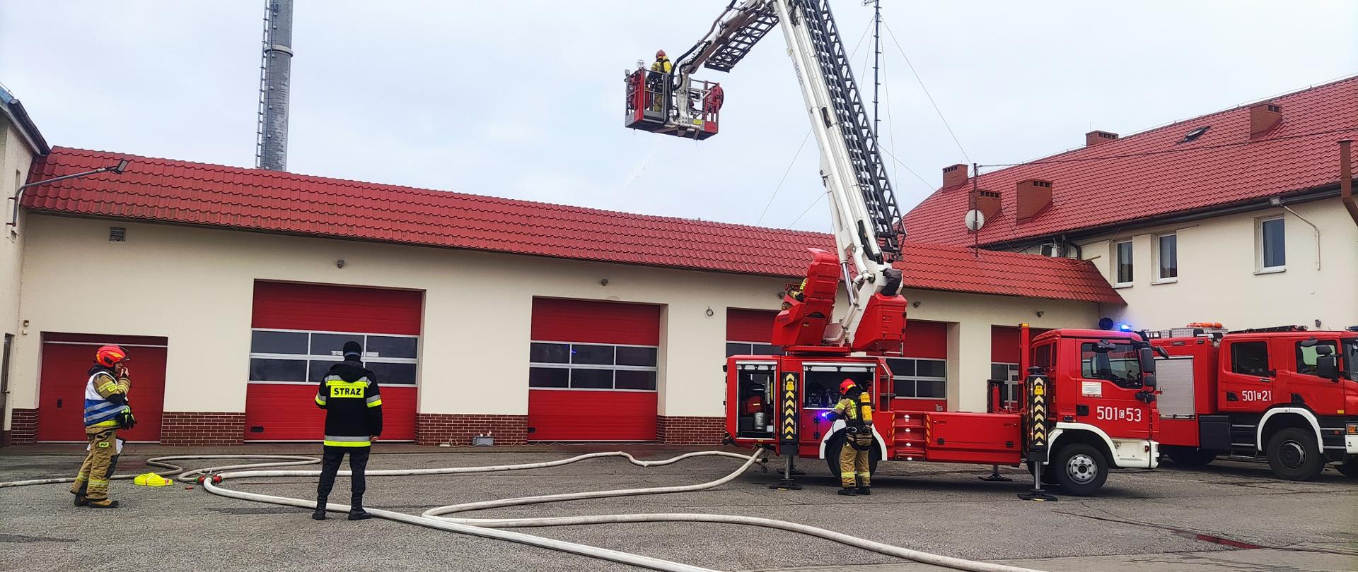Zdjęcie przedstawia teren Komendy Powiatowej Państwowej Straży Pożarnej, dwa wozy strażackie oraz strażaków podczas inspekcji gotowości operacyjnej