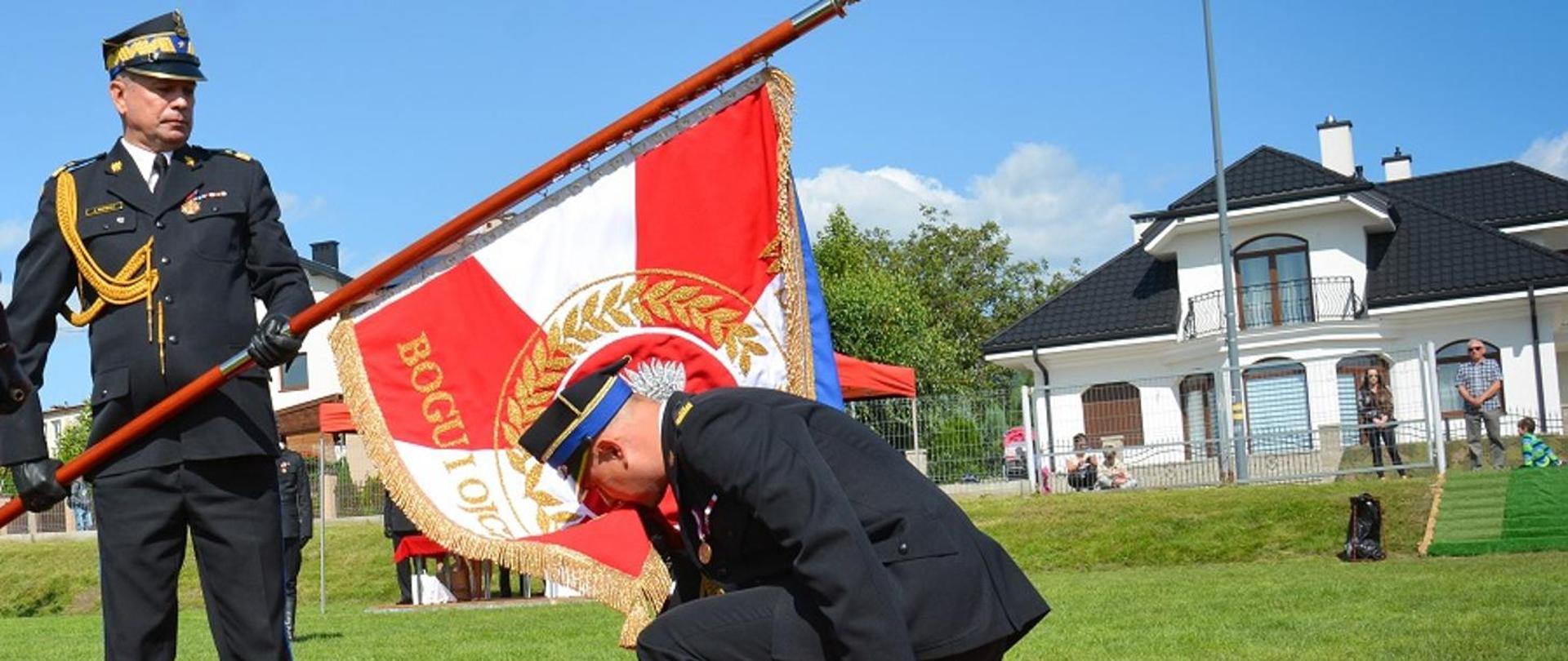 Plac apelowy uroczystości nadania sztandaru komendzie powiatowej Państwowej Straży Pożarnej w Mielcu. Na fotografii widać moment przekazania przez zastępcę komendanta głównego PSP nowo nadanego sztandaru komendzie powiatowej PSP w Mielcu. Na podeście okrytym czerwonym suknem stoi trzymając sztandar zastępca komendanta głównego. Przy sztandarze klęcząc na jedno kolano komendant powiatowy PSP w Mielcu całuje róg sztandaru. Strażacy ubrani są w mundury wyjściowe koloru granatowego. Głowy mają nakryte czapkami rogatywkami.