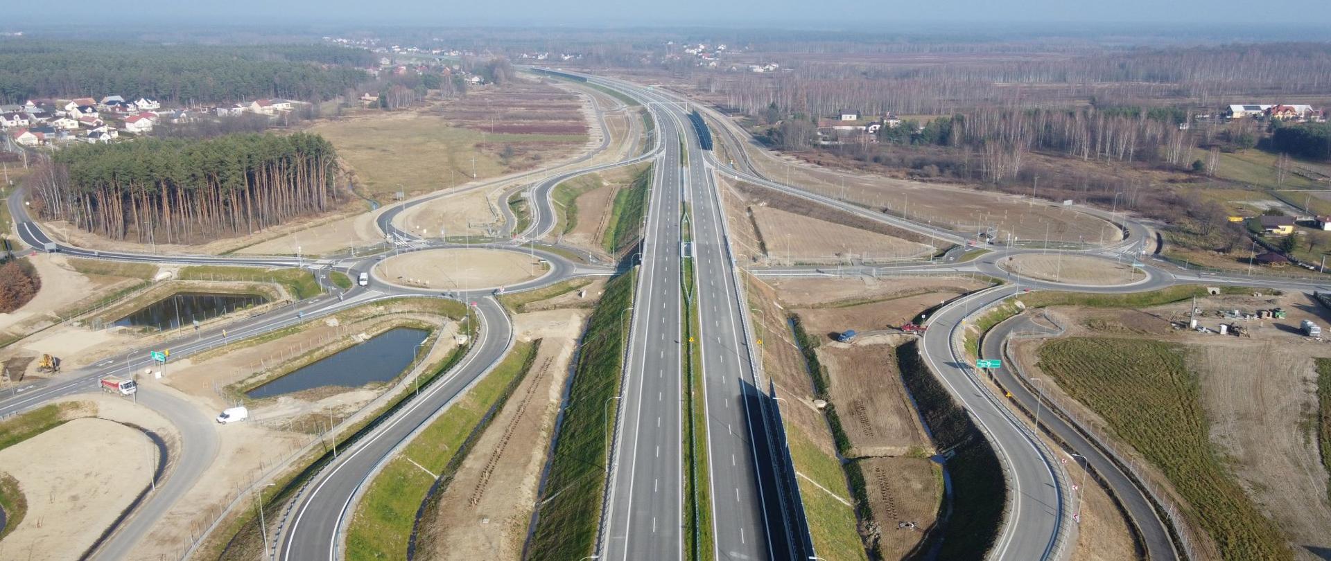 S19 węzeł Jeżowe w woj. podkarpackim, widok z powietrza na dwujezdniową drogę ekspresową w centrum kadru oraz odchodzące na lewo i prawo łącznice węzła. Po obu stronach drogi ekspresowej są duże ronda do których dochodzą łącznice węzła. 