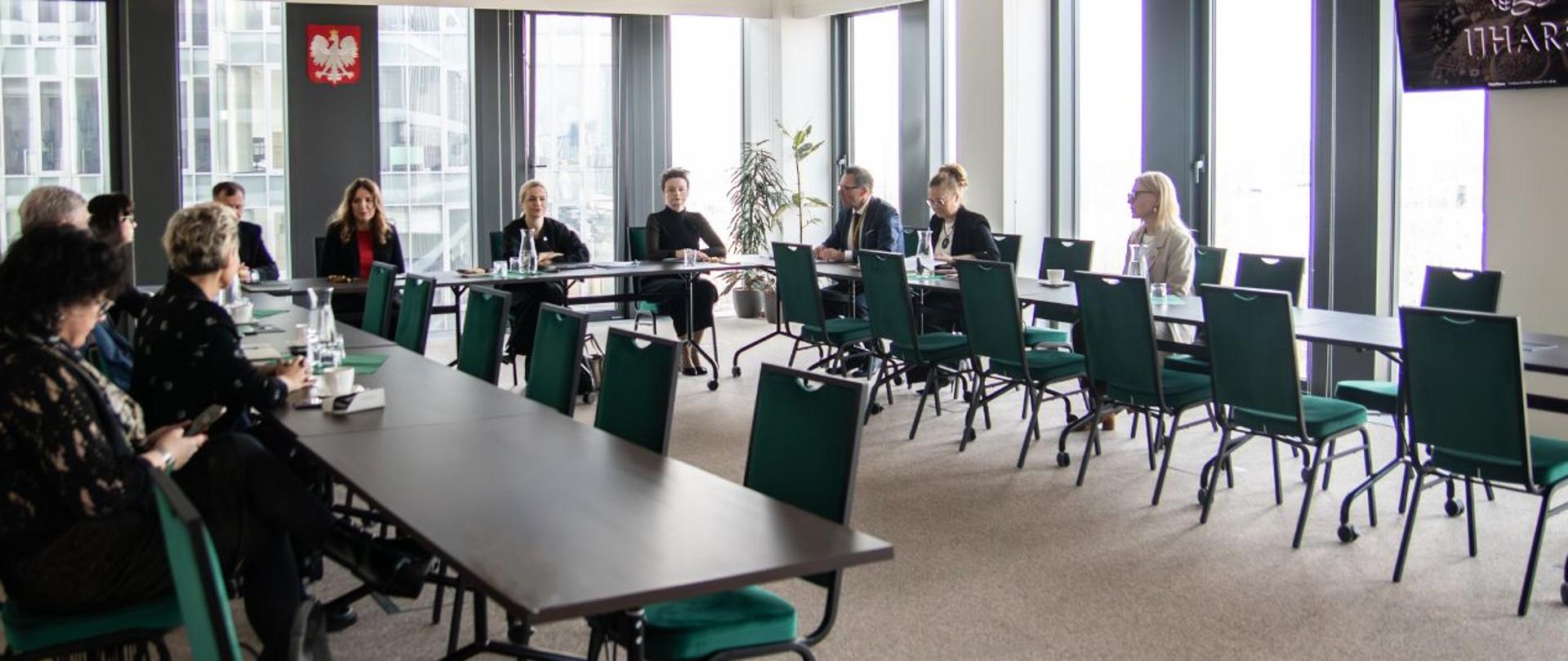 Sala konferencyjna z dużymi oknami, przy stołach ustawionych w podkowę siedzi kilka osób uczestniczących w spotkaniu; na ścianie widoczny jest herb Polski.