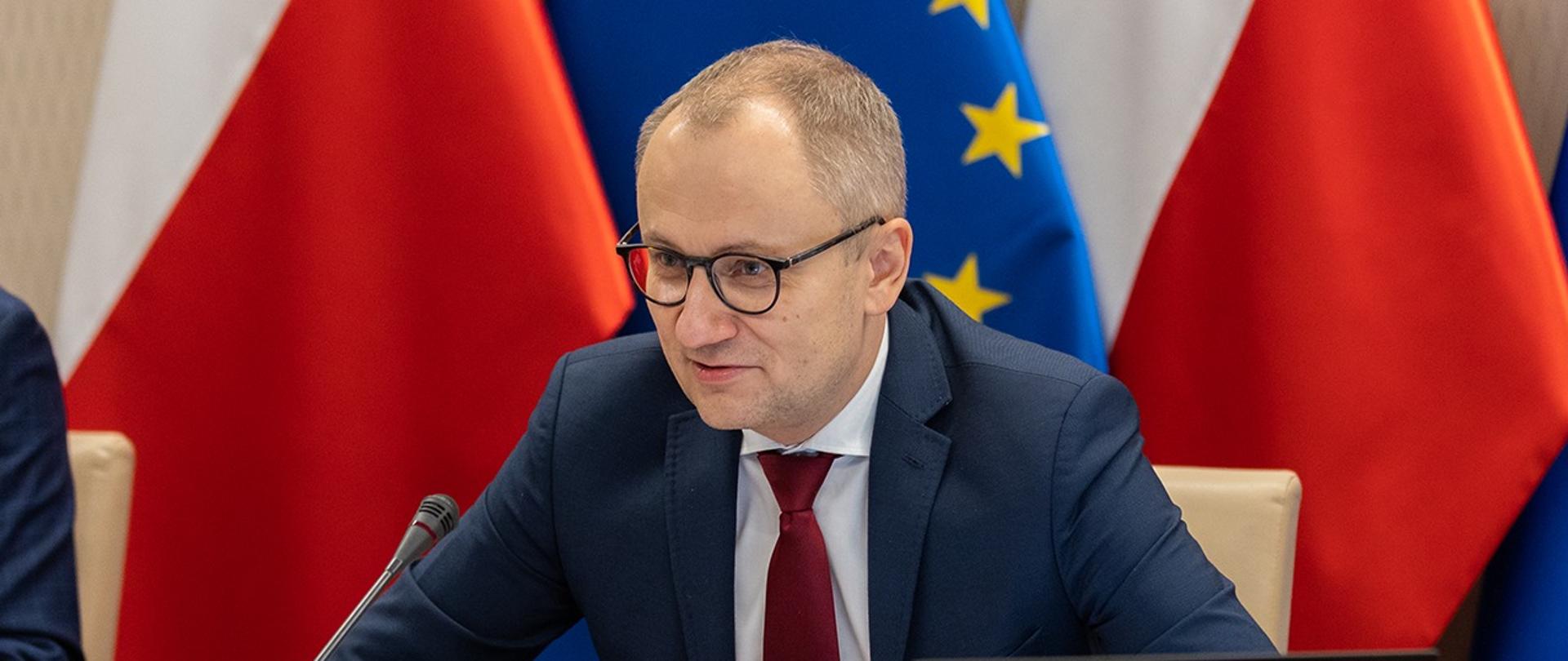 Wiceminister Błażej Poboży na tle flag Polski i Unii Europejskiej.
