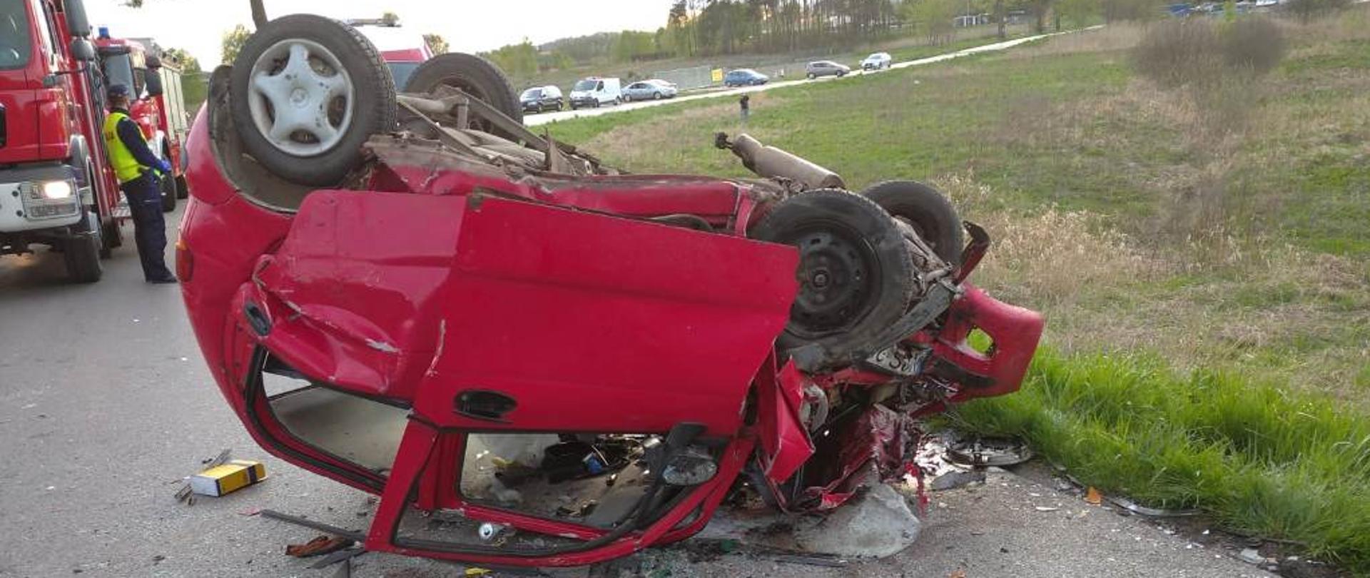 Zniszczony dachujący samochód po zderzeniu. W tle widać samochody ratowniczo-gaśnicze oraz policjanta zabezpieczającego miejsce zdarzenia.