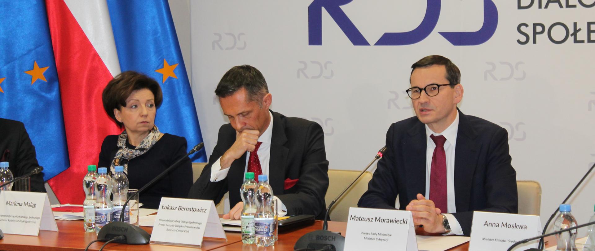 Na zdjęciu widnieje stół prezydialny za którym siedzi od lewej strony: Marlena Maląg - Minister Rodziny i Polityki Społecznej, Łukasz Bernatowicz - Przewodniczący rady Dialogu Społecznego oraz Premier Mateusz Morawiecki - Prezes Rady Ministrów. W tle siedzących baner z napisem Rada Dialogu Społecznego. Po lwewj stronie trzy flagi: UE, Polski i UE. 