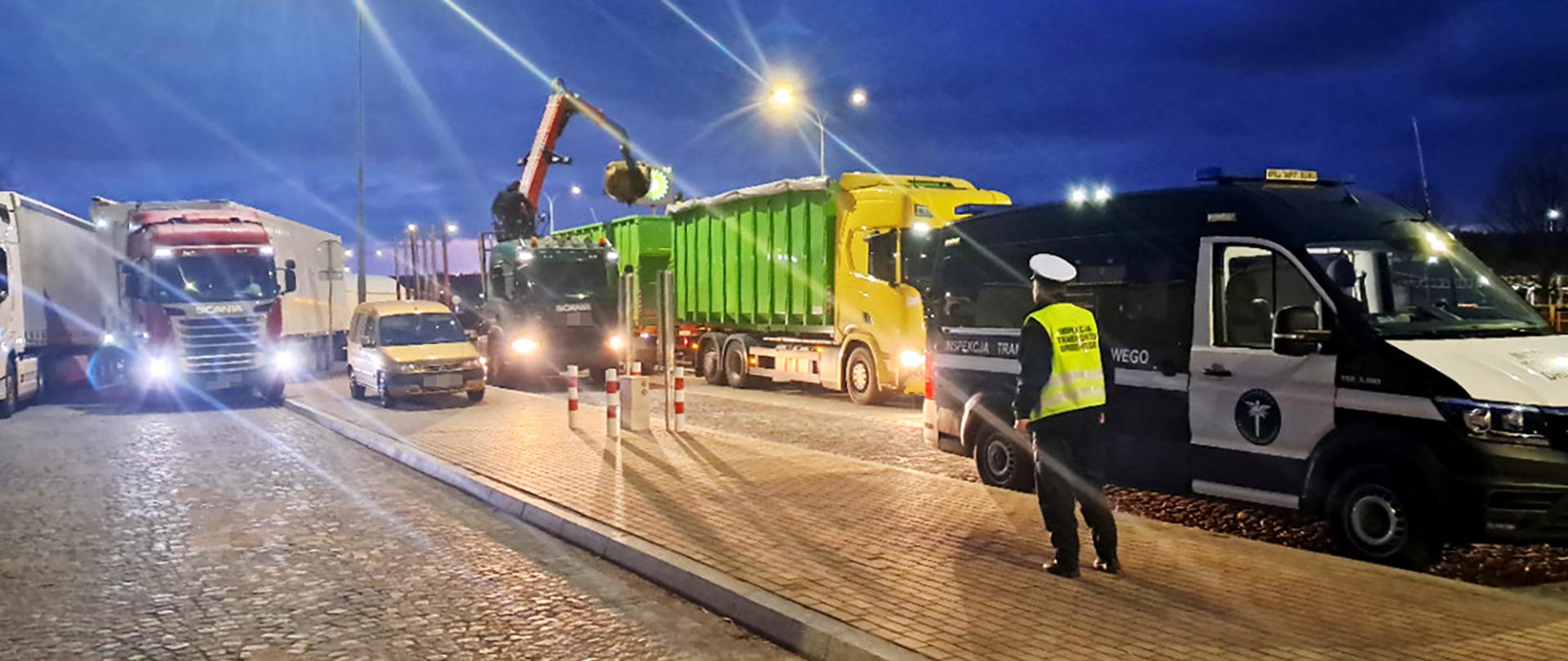 Nocna kontrola drogowa z udziałem inspektora w kamizelce odblaskowej stojącego obok oznakowanego pojazdu służbowego; przed nim ustawiony jest zestaw, z którego część ładunku jest przeładowywana koparką na inny pojazd ciężarowy. Pora nocna.