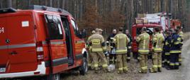 Na leśnej drodze zaparkowane są wozy strażackie, a przed nimi zgromadziła się liczna grupa strażaków OSP i PSP. Uczestnicy stoją w półkolu wokół stołu z przygotowanym sprzętem, wysłuchując instrukcji prowadzących. Scena przedstawia jedną ze stacji szkoleniowych podczas ćwiczeń, gdzie omawiane są zasady obsługi urządzeń i technik ratowniczych.