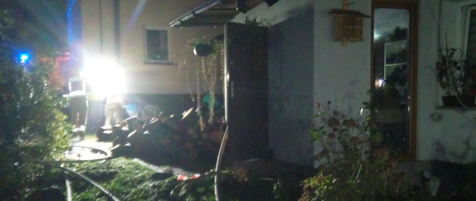 Zdjęcie przedstawia posesję na której doszło do pożaru. Z prawej strony budynek kotłowni – tam wprowadzone są węże gaśnicze. W głębi budynek oraz najaśnica oświetlająca miejsce działań.