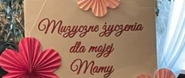 Zdjęcie kolorowe. Beżowa tabliczka z czerwonym napisem „Muzyczne życzenia dla mojej Mamy” ozdobiona trzema papierowymi kwiatkami w odcieniach różu. Dwa kwiatki są w kolorze ciemnoróżowym, a jeden jasnoróżowy. Tło stanowi roślina doniczkowa z zielonymi liśćmi.