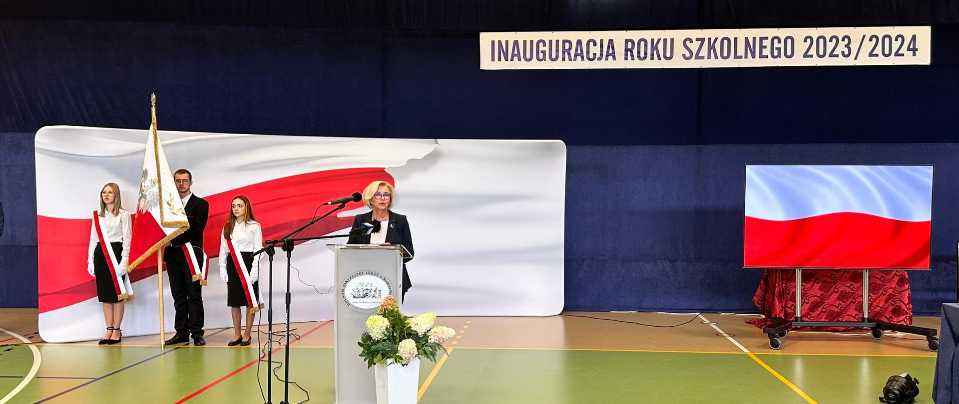 Na środku szkolnej sali gimnastycznej wiceminister Machałek stoi za mównicą i mówi do mikrofonu, za nią na tle polskiej flagi stoi troje uczniów ze sztandarem.