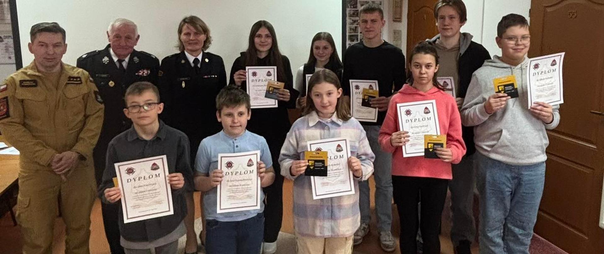 Na zdjęciu w dwóch rzędach stoją dzieci i młodzieży, laureaci konkursu oraz dorosłe osoby z czego trójka z nich jest w mundurach. Dzieci i młodzież trzymają w dłoniach dyplomy oraz nagrody.