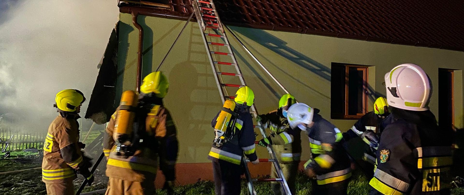 Strażacy przed budynkiem mieszkalnym, z lewej strony widoczne zadymienie, strażacy ustawiają drabinę