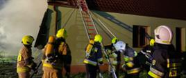Strażacy przed budynkiem mieszkalnym, z lewej strony widoczne zadymienie, strażacy ustawiają drabinę