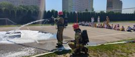Strażak z prądownica gaszący pożar tacy z benzyną obok zgromadzone dzieci na pokazie