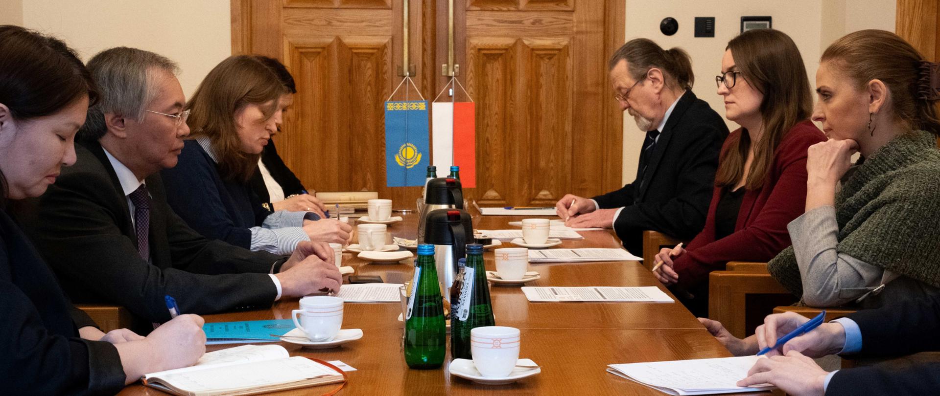 Po obu stronach stołu siedzą kobiety i mężczyźni. Na stole filiżanki i notatki. W tle flagi UE i Polski 