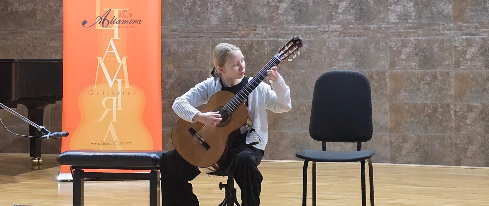 Kolorowe zdjęcie przedstawiające młodą gitarzystkę występującą na scenie. Dziewczyna siedzi na specjalnym taborecie, trzymając gitarę klasyczną w prawidłowej pozycji koncertowej (lewa noga na podnóżku). Jest ubrana w biały kardigan, czarną bluzkę i szerokie czarne spodnie. W tle widoczna jest ściana o fakturze szarego kamienia, na której wisi godło Polski. Po lewej stronie stoi pomarańczowy baner reklamowy "Altamira Guitarras", a na podłodze przed sceną znajdują się czarne głośniki sceniczne marki FBT.