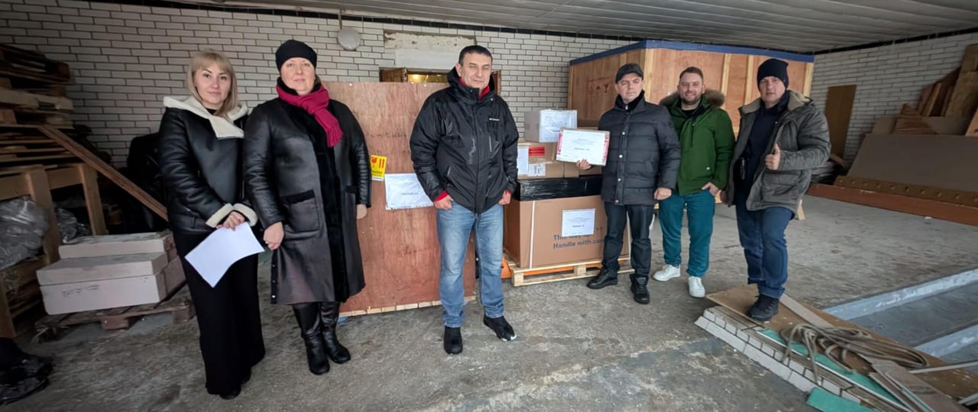 Grupa kobiet i mężczyzn stoi na tle palet z paczkami