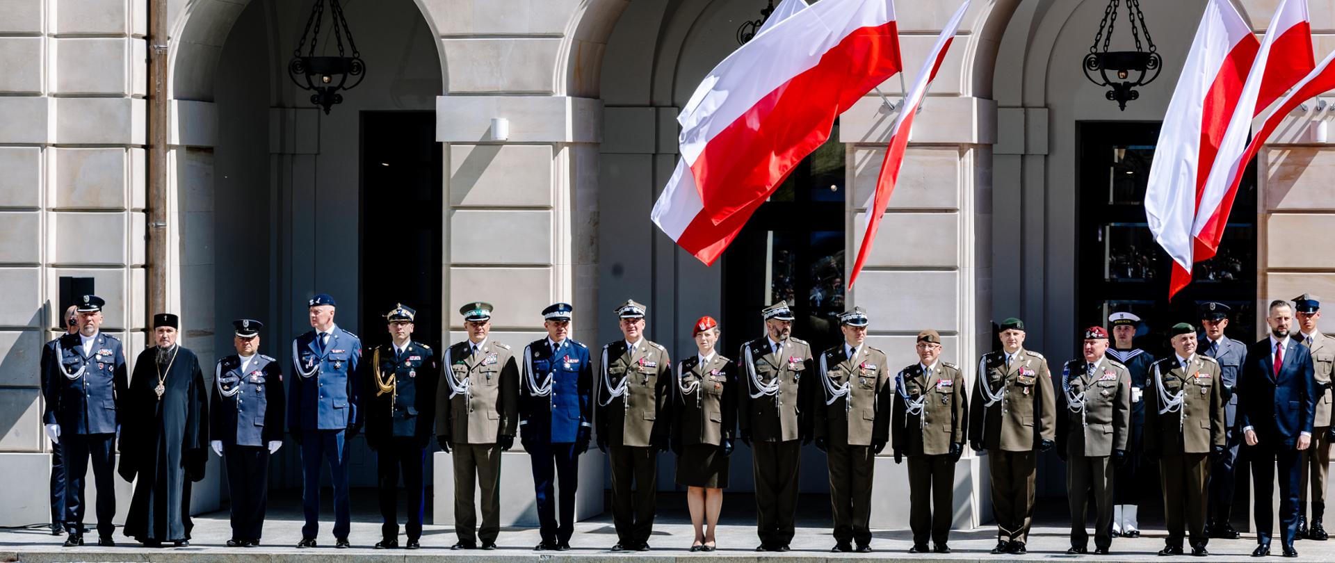 Przed arkadami pałacowego budynku stoi rząd przedstawicieli służb mundurowych, nad którymi powiewają biało-czerwone flagi.