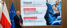 Wiceminister Gzik stoi na sali i mówi do mikrofonu na stojaku, za nim pod ścianą z drewnianych listewek flagi Polski i UE, na wbudowanym w ścianę ekranie napis Podwyżki dla nauczycieli.