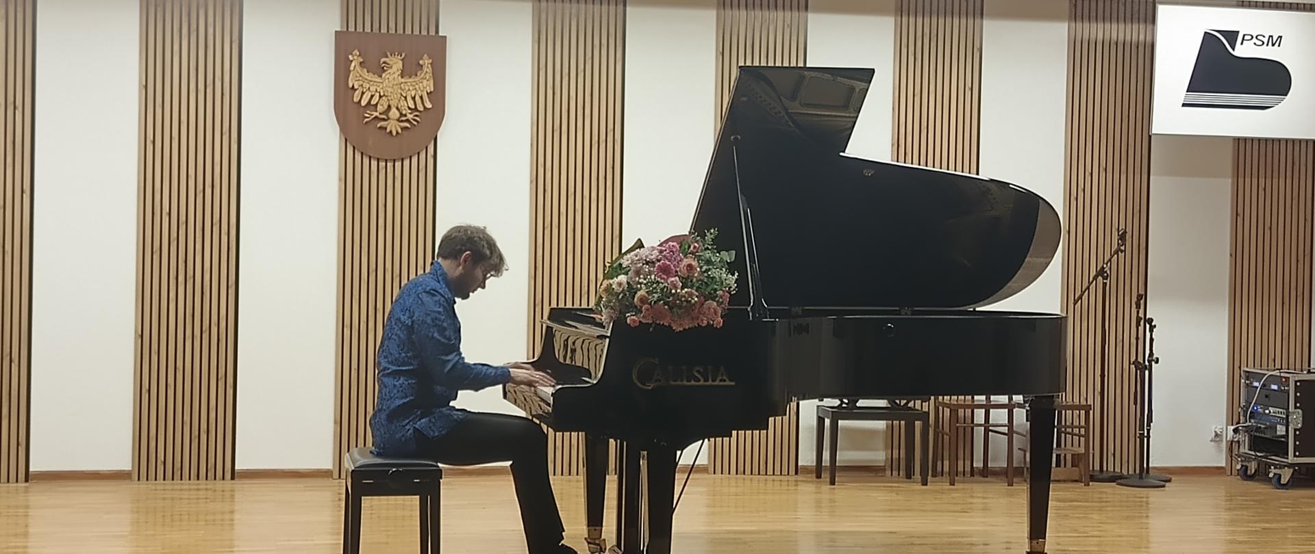 Na scenie stoi czarny fortepian, na nim leżą kwiaty, na fortepianie gra mężczyzna, w tle biała ściana z drewnianymi dekoracjami.