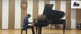 Na scenie stoi czarny fortepian, na nim leżą kwiaty, na fortepianie gra mężczyzna, w tle biała ściana z drewnianymi dekoracjami.