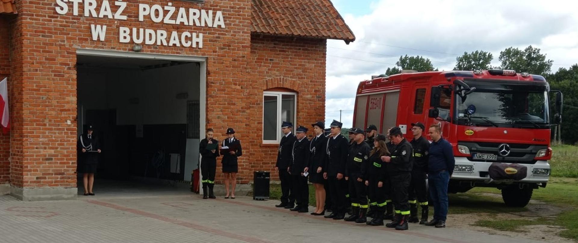 Druhowie OSP stoją przed remizą na bacznosć za nimi stoi samochód gaśniczy.