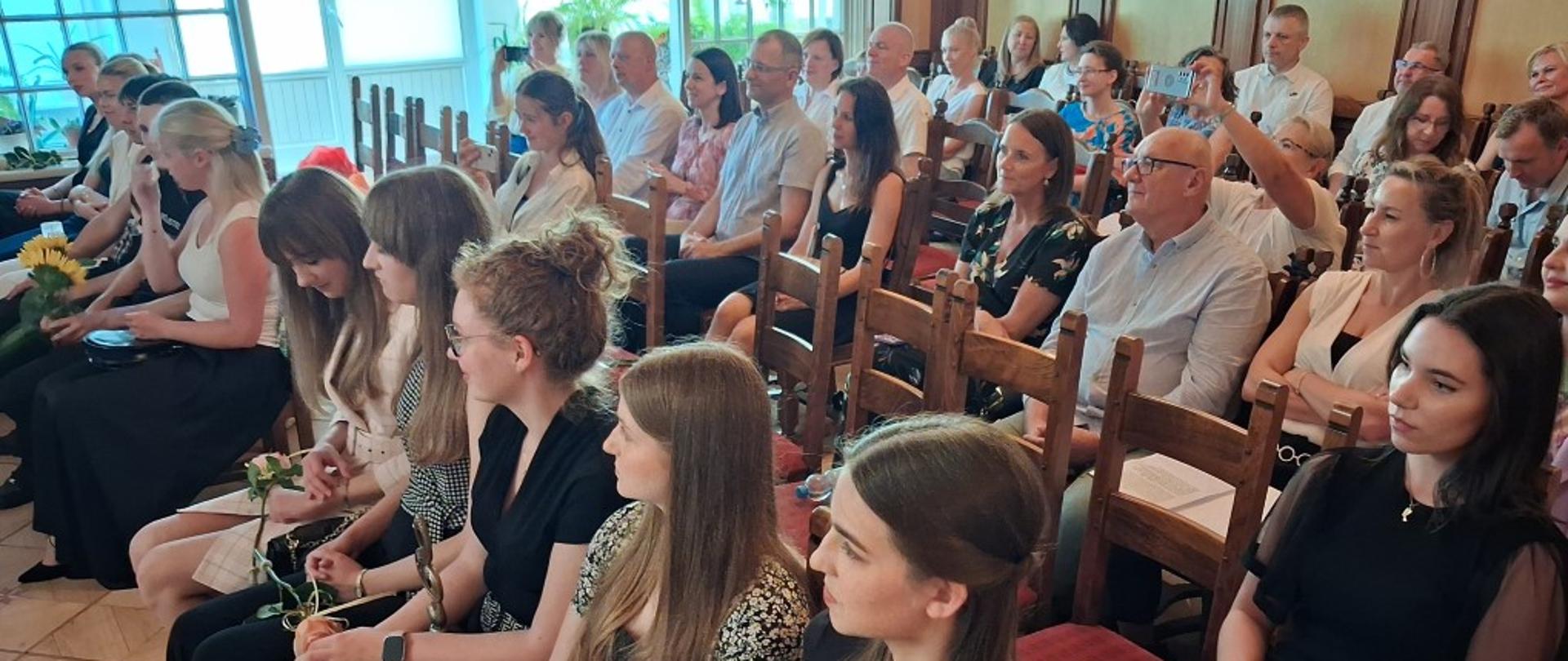 Zdjęcie kolorowe. Grupa osób siedząca na drewnianych krzesłach w rzędach w jasnej sali. W pierwszym rzędzie po prawej siedzą młode kobiety w ciemnych i wzorzystych strojach, jedna trzyma bukiet kwiatów. Za nimi widoczni są pozostali uczestnicy spotkania – kobiety i mężczyźni, z których część robi zdjęcia telefonami. Wszyscy patrzą w lewą stronę. W tle znajdują się duże okna, a wnętrze wykończone jest drewnianymi elementami.