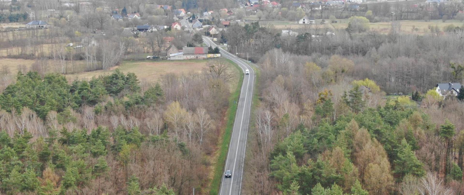 Widok z lotu ptaka na prosty odcinek drogi biegnącej przez las i tereny wiejskie, z kilkoma pojazdami jadącymi w obu kierunkach; w tle widoczne zabudowania i pola.