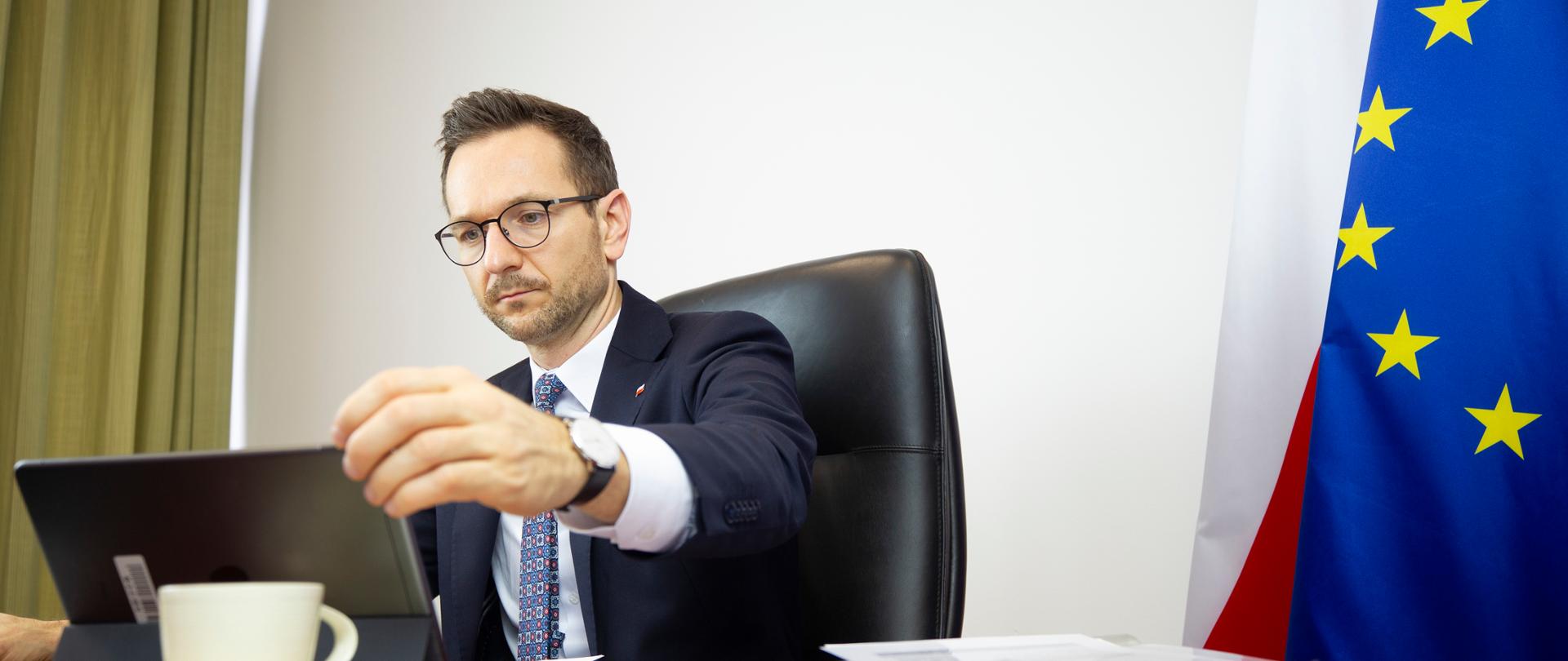 wiceminister Waldemar Buda patrzy w kierunku komputera z transmisją online, za nim flagi Polski i UE