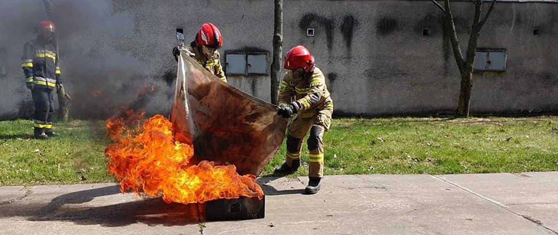 Na zdjęciu dwóch strażaków przy pomocy koca gaśniczego próbuje ugasić wysoki na około 1 m płomień. W tle stoi strazak, który przygląda się temu działaniu.