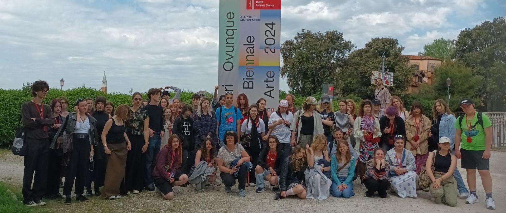 Na zdjęciu widzimy uczestników Artystycznej Wyprawy do Włoch - uczniowie oraz nauczyciele Państwowego Liceum Sztuk Plastycznych im. Antoniego Kenara w Zakopanem. Na drugim planie centralnie widzimy baner reklamowy informujący o 60 Biennale Sztuki odbywającym się w Wenecji. W tle widzimy krzewy i drzewa oraz zarys miasta.