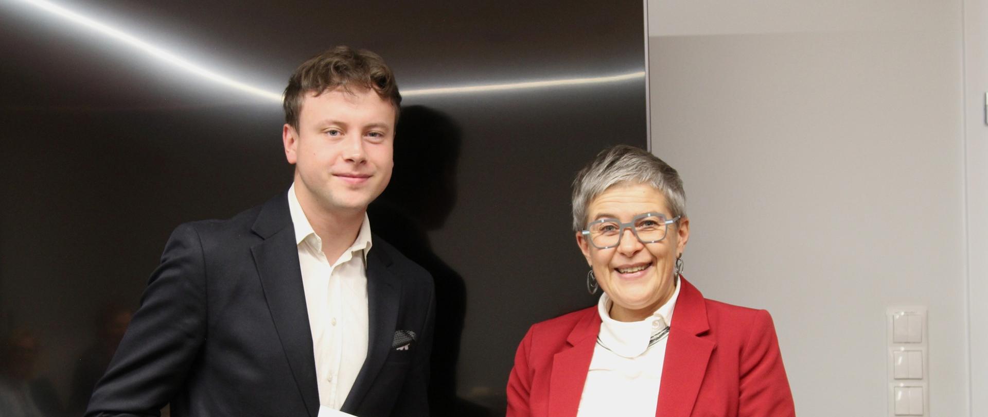 Wiceminister Mrówczyńska w czerwonej marynarce stoi obok mężczyzny w czarnej marynarce trzymającego białą teczkę z napisem Ministerstwo Nauki i Szkolnictwa Wyższego.