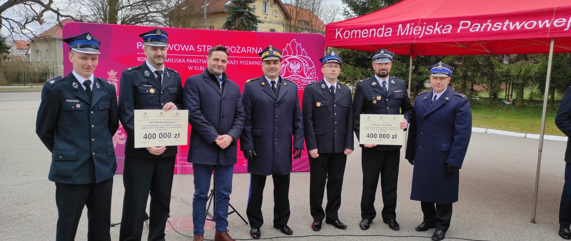 Na tle czerwonego baneru Państwowej Straży Pożarnej stoi sześciu strażaków strażacy w mundurach wyjściowych, w środku pomiędzy strażakami stoi Prezes Zarządu Wojewódzkiego Funduszu Ochrony Środowiska i Gospodarki Wodnej w Olsztynie.