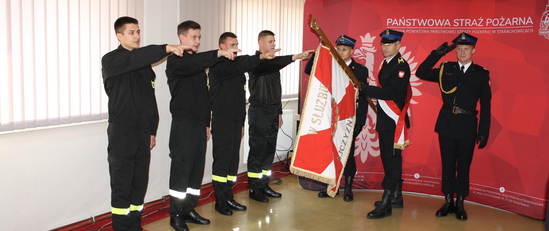 Z lewej strony czterech nowych funkcjonariuszy w czarnych ubraniach koszarowych stoją na baczność z wyciągniętymi prawymi rękoma. Z prawej strony poczet sztandarowy w mundurach galowych z sztandarem Komendy na tle czerwonej ścianki z symbolami narodowymi.